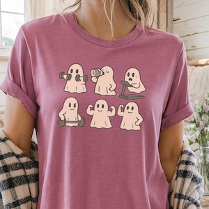 May include: A mauve-colored t-shirt featuring a graphic of six cartoon ghosts engaged in various gym activities. The ghosts are depicted lifting weights, using a treadmill, and flexing their muscles. The shirt has short sleeves and a relaxed fit.