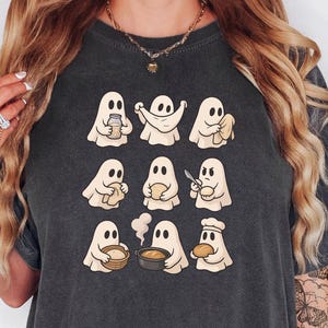 May include: Dark gray t-shirt featuring a grid of nine cartoon ghosts. Each ghost is depicted in a different cooking-related scenario, such as holding a jar, wrapping itself in a cloth, or holding a knife. The ghosts are light beige with black eyes.