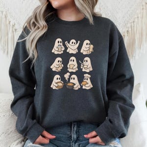 May include: Dark gray sweatshirt featuring a repeating pattern of cartoon ghosts in various cooking poses. The ghosts are beige with black eyes and are holding items like bread, bowls, and utensils. The sweatshirt is a casual, comfortable style.