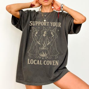 Może przedstawiać: Ciemnoszara koszulka z napisem "SUPPORT YOUR LOCAL COVEN" nad ilustracją trzech czarownic wok&oacute;ł kotła. Czarownice mają na sobie długie suknie i spiczaste kapelusze. Koszulka ma wygląd vintage.
