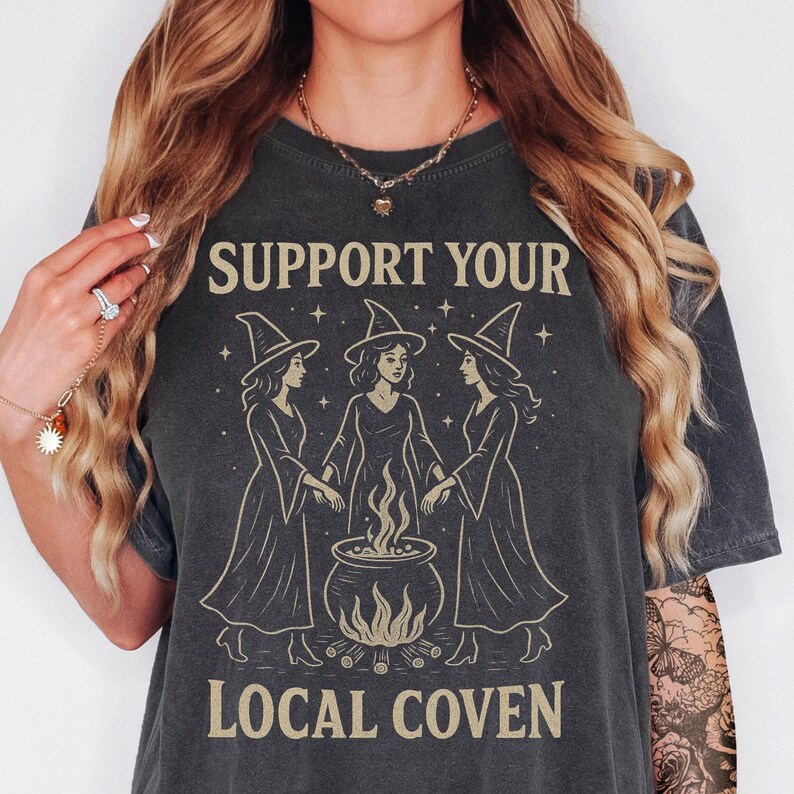 Może przedstawiać: Ciemnoszara koszulka z napisem "SUPPORT YOUR LOCAL COVEN". Koszulka przedstawia grafikę trzech czarownic w spiczastych kapeluszach stojących wok&oacute;ł kotła z płomieniami. Projekt jest w jasnobrązowym kolorze.