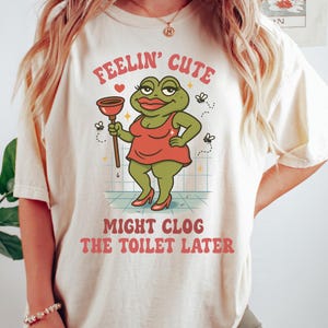 K&ouml;nnte beinhalten: Cremefarbenes T-Shirt mit einer Cartoon-Fr&ouml;schin in rotem Kleid und Abs&auml;tzen, die einen P&uuml;mpel h&auml;lt. Der Text lautet "FEELIN' CUTE" dar&uuml;ber und "MIGHT CLOG THE TOILET LATER" unter der Frosch-Illustration. Kleine Fliegen sind ebenfalls abgebildet.