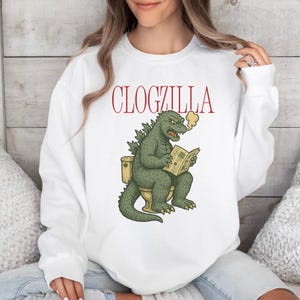 May include: White sweatshirt with the red text "CLOGZILLA" above a cartoon illustration of a green Godzilla-like creature sitting on a toilet, reading a newspaper, and exhaling smoke.