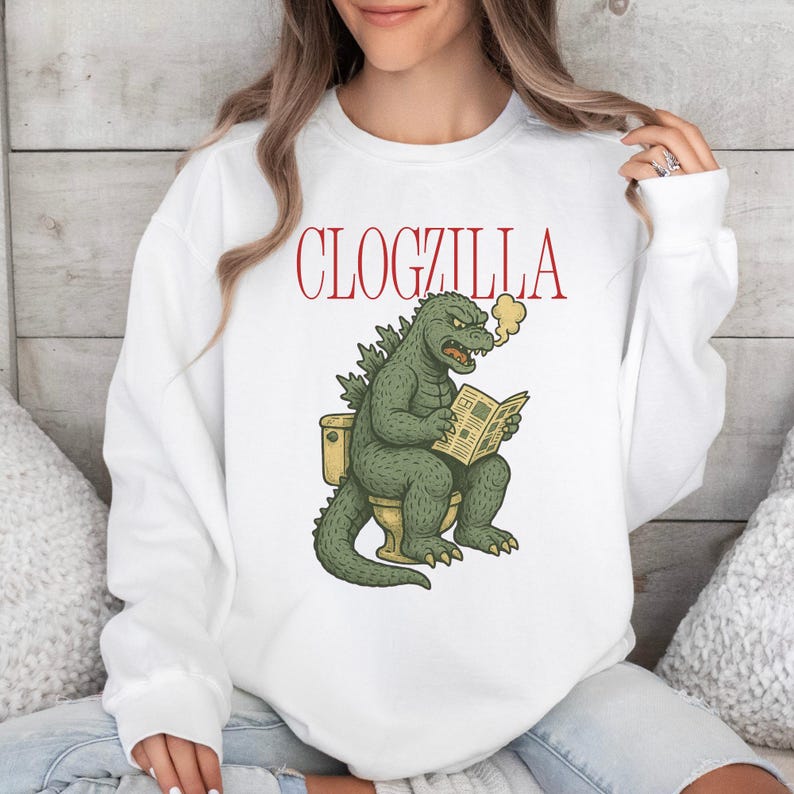 May include: White sweatshirt with the red text "CLOGZILLA" above a cartoon illustration of a green Godzilla-like creature sitting on a toilet, reading a newspaper, and exhaling smoke.