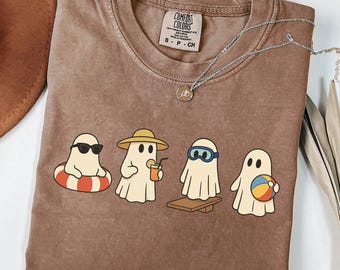 May include: Brown Comfort Colors t-shirt featuring a graphic of four cartoon ghosts enjoying summer activities. One ghost wears sunglasses and floats on a red and white inner tube. Another wears a hat and holds a drink. The shirt has a small tag.