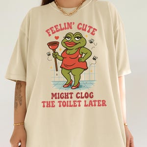 K&ouml;nnte beinhalten: Beiges T-Shirt mit einer Cartoon-Fr&ouml;schin in rotem Kleid, die einen P&uuml;mpel h&auml;lt. Der Text "FEELIN' CUTE" und "MIGHT CLOG THE TOILET LATER" ist &uuml;ber und unter der Frosch-Illustration aufgedruckt.