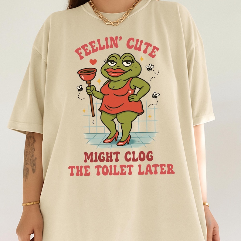 K&ouml;nnte beinhalten: Beiges T-Shirt mit einer Cartoon-Fr&ouml;schin in rotem Kleid, die einen P&uuml;mpel h&auml;lt. Der Text "FEELIN' CUTE" und "MIGHT CLOG THE TOILET LATER" ist &uuml;ber und unter der Frosch-Illustration aufgedruckt.
