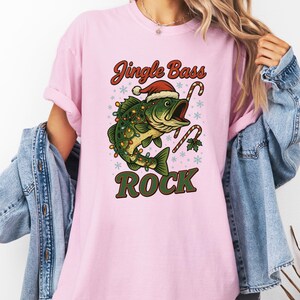 Pu&ograve; includere: T-shirt rosa con grafica a tema natalizio. Il design presenta un pesce verde con cappello di Babbo Natale e luci natalizie, con bastoncini di zucchero e le parole "Jingle Bass Rock".