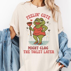 K&ouml;nnte beinhalten: Cremefarbenes T-Shirt mit einer Cartoon-Fr&ouml;schin in rotem Kleid und Abs&auml;tzen, die einen P&uuml;mpel h&auml;lt. Der Text "FEELIN' CUTE" und "MIGHT CLOG THE TOILET LATER" ist &uuml;ber und unter dem Frosch aufgedruckt. Das Design beinhaltet kleine fliegende Insekten.