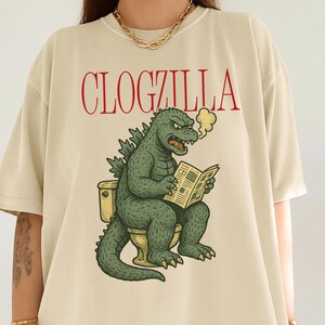 May include: Beige t-shirt with the red text "CLOGZILLA". The graphic shows a green Godzilla-like creature sitting on a toilet, reading a newspaper, and exhaling smoke. The design is a humorous parody.