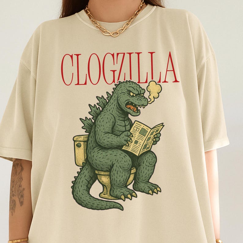May include: Beige t-shirt with the red text "CLOGZILLA". The graphic shows a green Godzilla-like creature sitting on a toilet, reading a newspaper, and exhaling smoke. The design is a humorous parody.