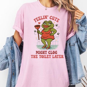 K&ouml;nnte beinhalten: Hellrosa T-Shirt mit dem Text "Feelin' Cute" und "Might Clog The Toilet Later." Das Design zeigt einen Cartoon-Frosch in einem roten Kleid, der einen P&uuml;mpel h&auml;lt und rote High Heels tr&auml;gt.