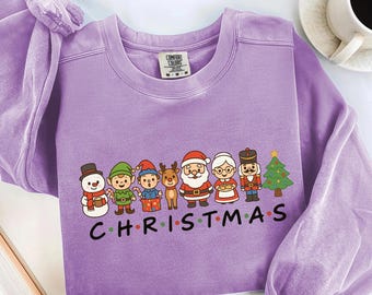 May include: A light purple sweatshirt featuring a festive graphic of Christmas characters, including a snowman, elves, Santa Claus, and a reindeer. The word "CHRISTMAS" is printed in black block letters below the characters. The sweatshirt has a crew neck and long sleeves.