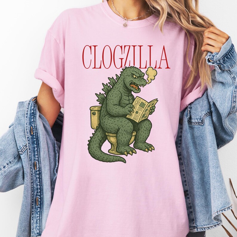 May include: Pink t-shirt with the text "CLOGZILLA" in red. The graphic features a green Godzilla-like creature sitting on a toilet, reading a newspaper, with steam rising from its mouth.