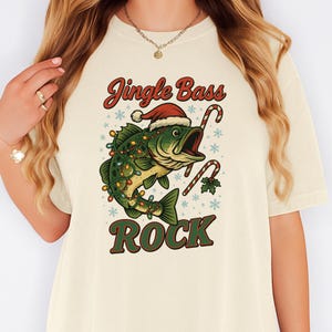 Pu&ograve; includere: T-shirt color crema con grafica natalizia. Presenta un pesce verde con cappello di Babbo Natale e luci natalizie, con le parole "Jingle Bass Rock". Bastoncini di zucchero e fiocchi di neve completano il design festivo.