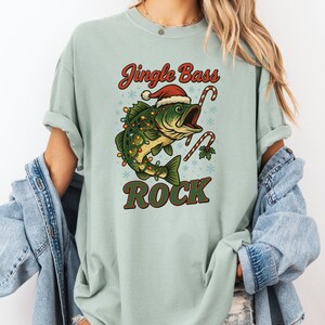 Pu&ograve; includere: T-shirt verde chiaro con grafica a tema natalizio. Il design include un pesce verde con cappello di Babbo Natale e luci natalizie, con bastoncini di zucchero e la scritta "Jingle Bass Rock".