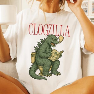 May include: White t-shirt with the text "CLOGZILLA" in red. The shirt features a green Godzilla-like creature sitting on a toilet, reading a newspaper, with steam coming from its mouth. The design is a humorous take on the classic monster.