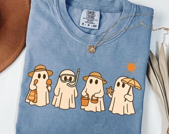 May include: A blue Comfort Colors t-shirt featuring a graphic of four cartoon ghosts at the beach. Each ghost is wearing a hat and holding beach items like ice cream, a snorkel, a bucket, and an umbrella. A gold necklace is draped across the shirt.