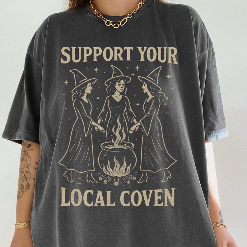 Może przedstawiać: Ciemnoszara koszulka z napisem "SUPPORT YOUR LOCAL COVEN". Grafika przedstawia trzy czarownice w długich szatach i spiczastych kapeluszach, wok&oacute;ł kotła z płomieniami. Projekt jest w stylu vintage.