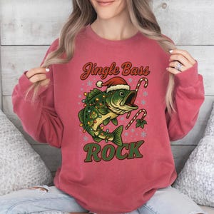 Pu&ograve; includere: Una felpa rosa-rossa con una grafica raffigurante un pesce con un cappello di Babbo Natale e luci natalizie. Il testo "Jingle Bass Rock" &egrave; sopra e sotto il pesce. Bastoncini di zucchero e fiocchi di neve completano il design natalizio.