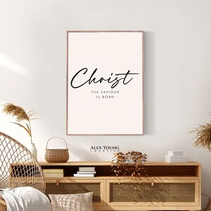 May include: A framed print with the word "Christ" in elegant script, with the text "The Saviour Is Born" below. The print has a light pink background and a thin, brown frame. The print is displayed above a wooden cabinet.