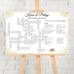Peut inclure: Un panneau blanc sur un chevalet avec un jeu de mots croisés pour un 50e anniversaire de mariage. Le panneau présente des accents floraux dorés et les noms "Karen & Philipp". Le texte "How well do you know Karen and Philipp?" est également visible.