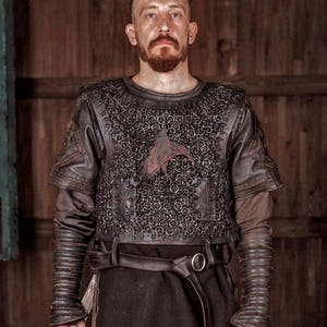 May include: A dark brown leather and chainmail armor vest featuring a central bird emblem, worn over a long-sleeved brown tunic with ribbed leather forearm guards and a wide leather belt with a metal buckle, creating a layered protective garment.