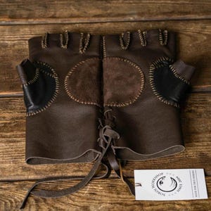 Pode incluir: Um par de luvas de couro castanho escuro sem dedos com um remendo circular de camurça no dorso da mão, costuras decorativas nas bordas e um fecho de atacadores no pulso. Uma etiqueta retangular branca contém o texto SOKOL WORKSHOP.