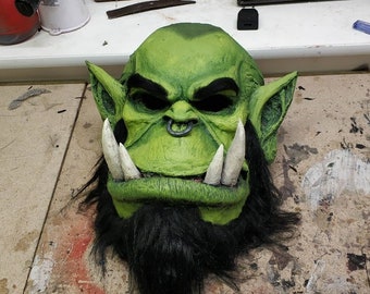 May include: A green orc mask with large white tusks, black eyebrows, and a black beard. The mask has a metal ring in the nose and is made of a textured material. The mask is likely for cosplay or costume use.