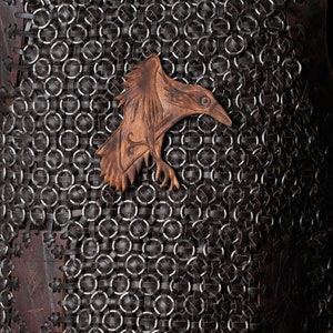 May include: A dark brown leather vest featuring a complex pattern of interlocking metal rings and leather strips. A carved leather raven emblem is centered on the chest, showcasing detailed feather textures and a sharp beak in a lighter tan tone.