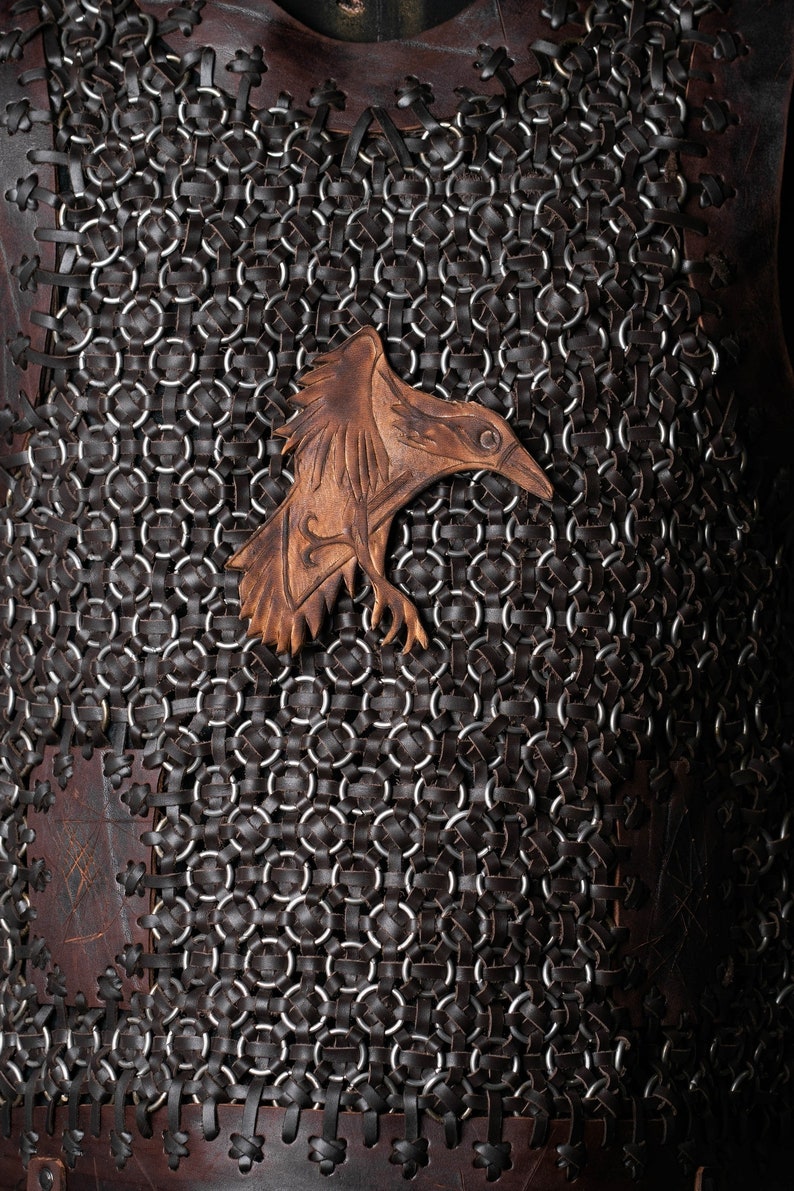 May include: A dark brown leather vest featuring a complex pattern of interlocking metal rings and leather strips. A carved leather raven emblem is centered on the chest, showcasing detailed feather textures and a sharp beak in a lighter tan tone.