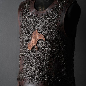 May include: A sleeveless vest constructed from dark brown leather panels and interconnected metal rings. A carved leather raven emblem is attached to the center of the chest. The garment features leather lacing along the edges and sides.