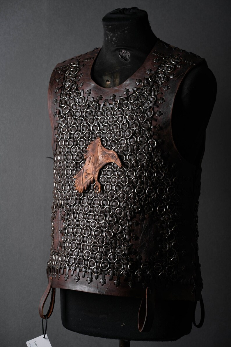 May include: A sleeveless vest constructed from dark brown leather panels and interconnected metal rings. A carved leather raven emblem is attached to the center of the chest. The garment features leather lacing along the edges and sides.