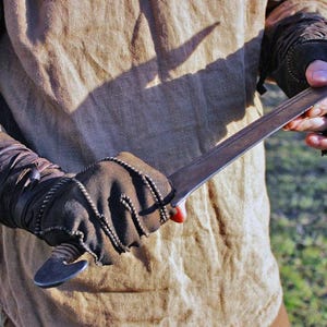 May include: A metal sword with a dark, weathered blade and a curved pommel, held by hands wearing brown fingerless leather gloves with decorative stitching and wrap-around wrist details, worn over a tan linen long-sleeved tunic.