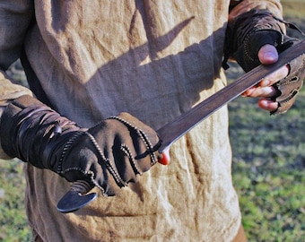 May include: A metal sword with a dark, weathered blade and a curved pommel, held by hands wearing brown fingerless leather gloves with decorative stitching and wrap-around wrist details, worn over a tan linen long-sleeved tunic.