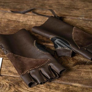 May include: A pair of fingerless dark brown leather gloves featuring suede patches, visible contrast stitching, and leather lacing. A white rectangular tag is attached, displaying the text Sokol Workshop Since 2006 Made For True Heroes.