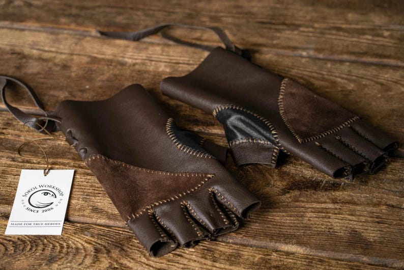 May include: A pair of fingerless dark brown leather gloves featuring suede patches, visible contrast stitching, and leather lacing. A white rectangular tag is attached, displaying the text Sokol Workshop Since 2006 Made For True Heroes.