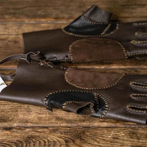 May include: A pair of fingerless dark brown leather gloves featuring contrasting suede patches, visible whipstitch detailing, and a lace-up closure at the wrist. A white rectangular tag attached to the gloves displays the text Sokol Workshop Since 2006.