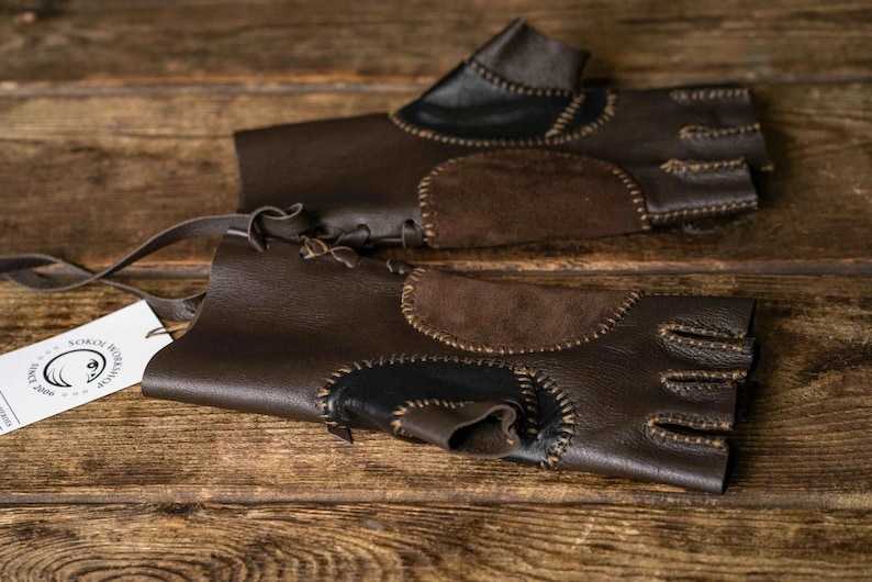 May include: A pair of fingerless dark brown leather gloves featuring contrasting suede patches, visible whipstitch detailing, and a lace-up closure at the wrist. A white rectangular tag attached to the gloves displays the text Sokol Workshop Since 2006.