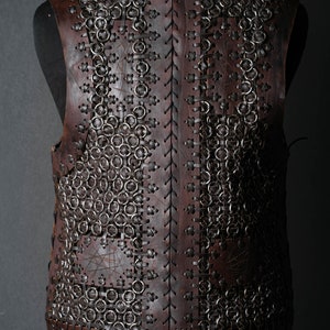 May include: A sleeveless vest constructed from dark brown leather panels and interconnected metal rings. The leather pieces are joined by thin leather lacing in a cross-stitch pattern, creating a structured garment with a visible texture.