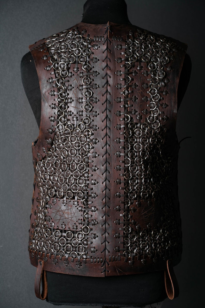 May include: A sleeveless vest constructed from dark brown leather panels and interconnected metal rings. The leather pieces are joined by thin leather lacing in a cross-stitch pattern, creating a structured garment with a visible texture.