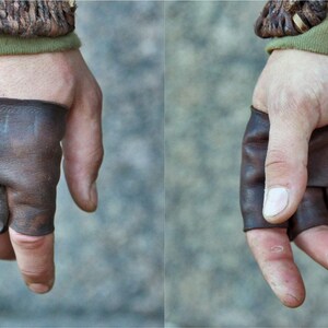 May include: A pair of fingerless brown leather gloves featuring brass-colored metal studs along the knuckles and faint, hand-drawn runic symbols on the back of the hand. The gloves are secured with leather lacing at the wrist area.