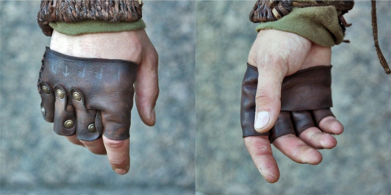 May include: A pair of fingerless brown leather gloves featuring brass-colored metal studs along the knuckles and faint, hand-drawn runic symbols on the back of the hand. The gloves are secured with leather lacing at the wrist area.