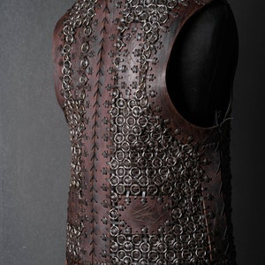 May include: A dark brown leather vest featuring a complex construction of interlocking metal rings secured with leather lacing. The garment displays a textured surface with visible stitching patterns and vertical leather panels connecting the chainmail sections.