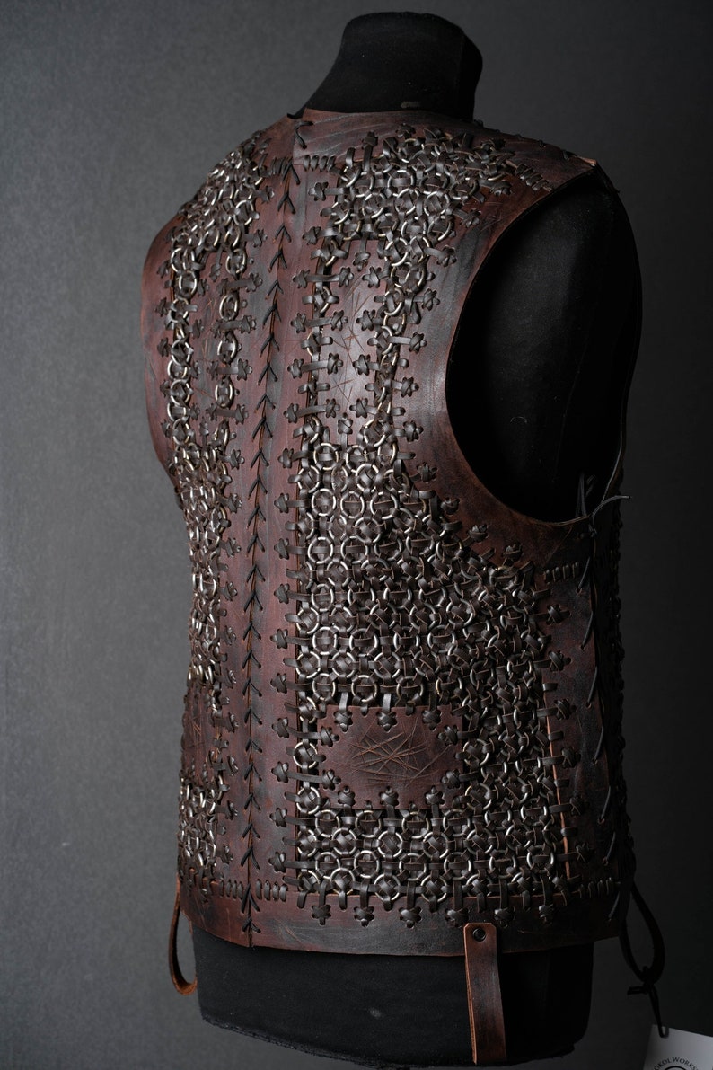 May include: A dark brown leather vest featuring a complex construction of interlocking metal rings secured with leather lacing. The garment displays a textured surface with visible stitching patterns and vertical leather panels connecting the chainmail sections.