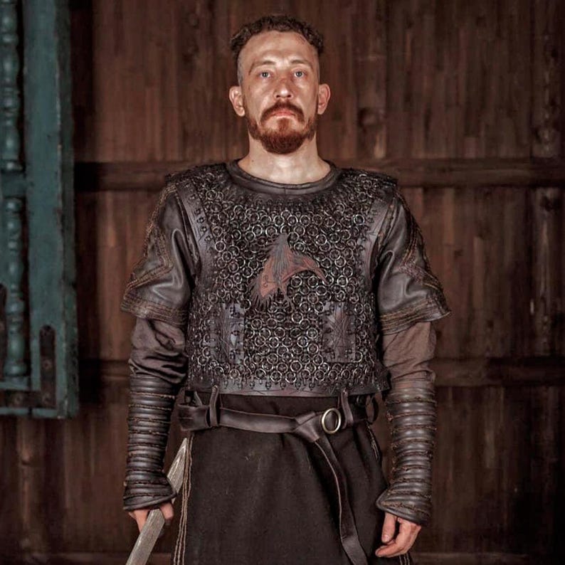 May include: A dark brown leather and chainmail armor vest featuring a central bird emblem, worn over a long-sleeved brown tunic with ribbed leather forearm guards and a wide leather belt with a metal buckle, creating a layered protective garment.