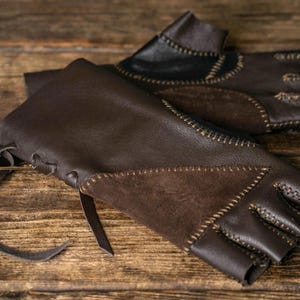 May include: A pair of fingerless gloves crafted from dark brown leather and suede panels. The gloves feature visible decorative stitching along the seams, a lace-up closure with leather ties at the wrist, and an open-finger design for dexterity.