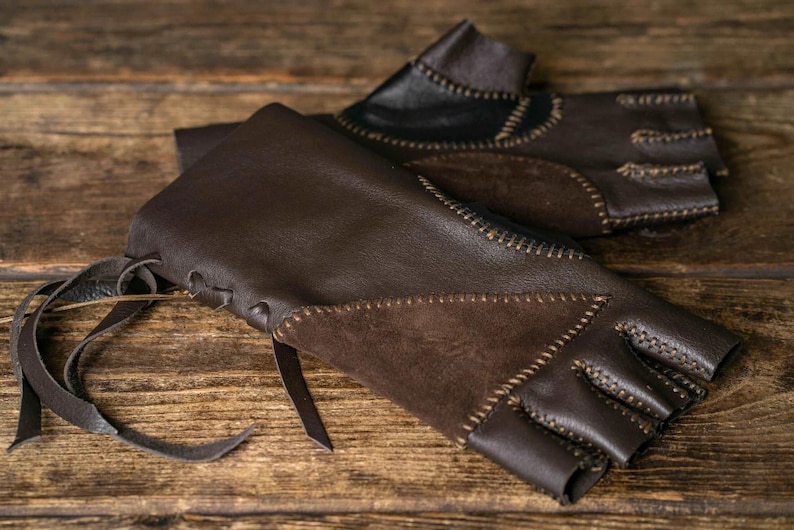 May include: A pair of fingerless gloves crafted from dark brown leather and suede panels. The gloves feature visible decorative stitching along the seams, a lace-up closure with leather ties at the wrist, and an open-finger design for dexterity.