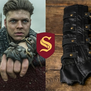 May include: A black leather forearm bracer featuring a series of adjustable straps with brass-colored metal buckles and rivets. The cuff is constructed with visible stitched seams along the edges and a tapered design at the wrist area.