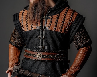 May include: A black hooded tunic featuring tan leather chest panels with black lacing, tan leather arm guards, and a wide brown leather belt with a circular metal buckle. A dark metal axe with a wrapped handle is held horizontally across the waist.
